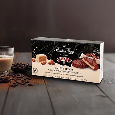 Verpackung der Anthon Berg Baileys Treats – Milchschokolade überzogenes Marzipan, verziert mit Mandeln, Schokolade und einem Glas Baileys im Hintergrund auf einem rustikalen Holztisch. Die Verpackung zeigt die Pralinen, eine aufgeschnittene Variante mit sichtbarer Marzipanfüllung, sowie ein Stück Marzipan und ganze Mandeln.