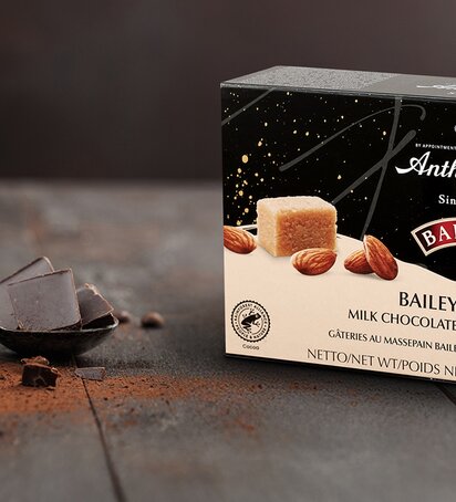 Verpackung der Anthon Berg Baileys Treats – Milchschokolade überzogenes Marzipan, verziert mit Mandeln, Schokolade und einem Glas Baileys im Hintergrund auf einem rustikalen Holztisch. Die Verpackung zeigt die Pralinen, eine aufgeschnittene Variante mit sichtbarer Marzipanfüllung, sowie ein Stück Marzipan und ganze Mandeln.
